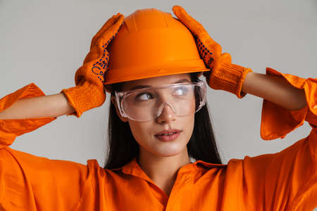 Young brunette woman in safety glasses and helmet holding her head isolated over grey background