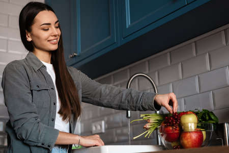 Happy mid aged brunette woman washing fruits and vegetables in a sink in the kitchen at home
