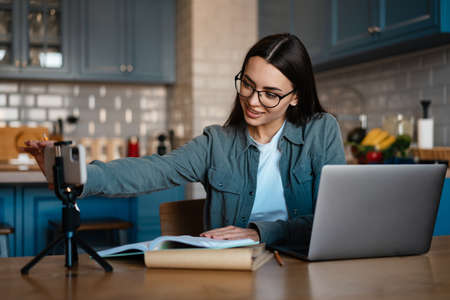 Smiling woman taking selfie footage on cellphone while working with laptop at home kitchen