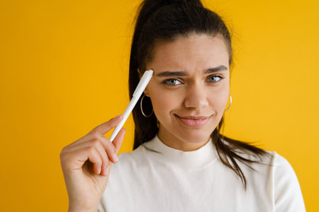 Young brunette woman smiling while posing with pen isolated over yellow background