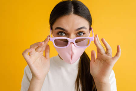 Young brunette woman in face mask and sunglasses looking at camera isolated over yellow backgroundの写真素材