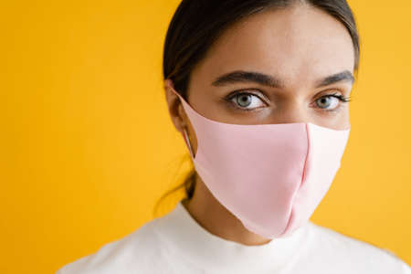 Young brunette woman in face mask posing and looking at camera isolated over yellow backgroundの写真素材