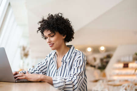 Beautiful happy curly woman working with laptop while sitting in cafeの写真素材