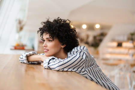 Beautiful pleased curly woman looking out window while sitting in cafeの写真素材
