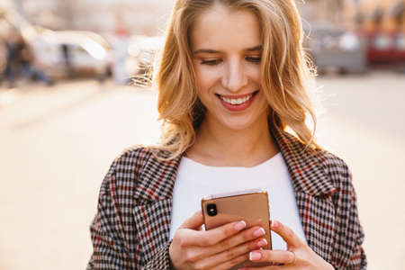 Smiling young blonde woman using mobile phone walking on a city streetの写真素材