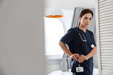 Focused mid woman doctor in medical suit fastening badge while standing at homeの写真素材