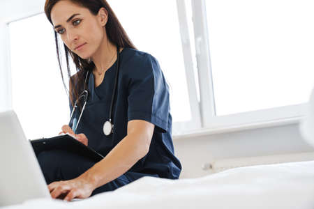 Focused woman doctor with stethoscope writing while working with laptop at home
