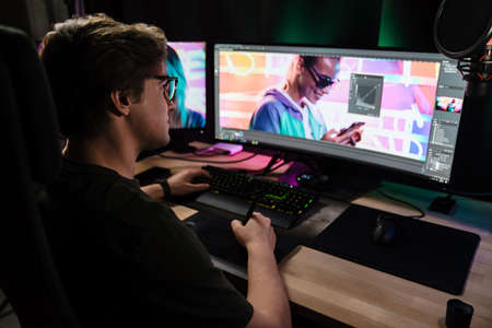 Back view of a young white man videographer working on a project with a touchpad on computer sitting...