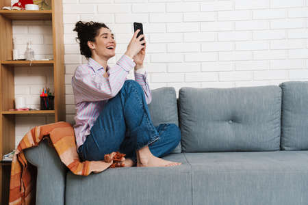 The curly happy woman sitting with legs on the sofa and looking at the phone in the light roomの写真素材