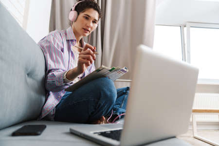 Portrait of a mid aged young woman in headphones studying while sitting taking notes at a couch at...