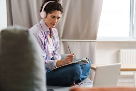 Portrait of a mid aged young woman in headphones studying while sitting taking notes at a couch at...