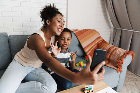 Black smiling woman taking selfie photo on cellphone while hugging her son at homeの写真素材