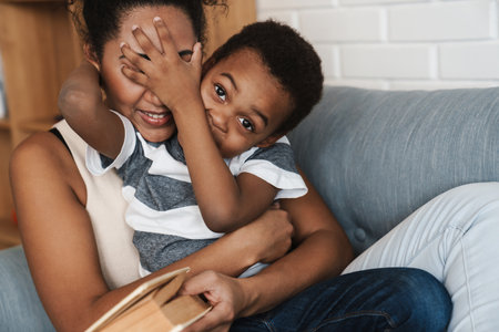 Black smiling mother and son making fun and reading book on sofa at homeの写真素材