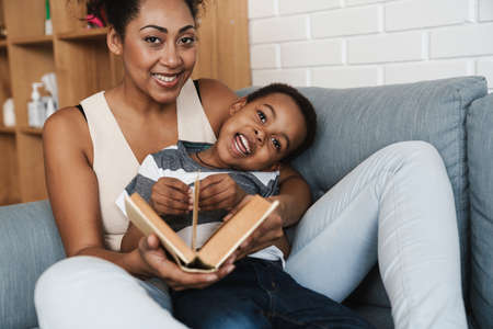 Black smiling mother and son reading book while sitting on sofa at homeの写真素材