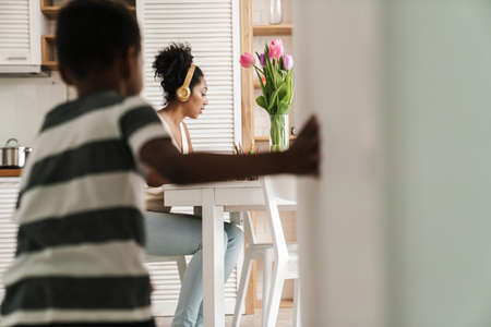 Black boy running and making fun while her mother working at table in homeの写真素材