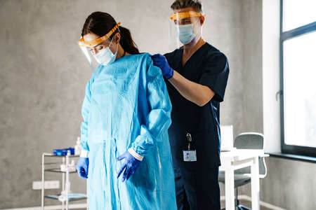 Two medical workers dressing up in uniform standing indoors, wearing robes and masksの写真素材