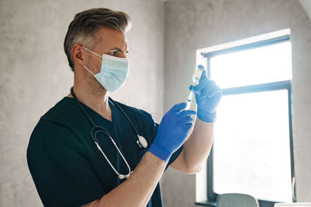 Mid aged male doctor wearing protective mask holding a syringe with vaccine