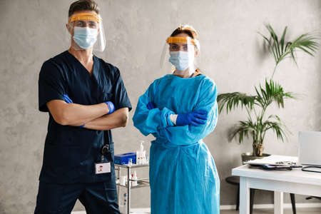 Two medical workers dressed in uniform standing indoors, wearing robes and masks, looking at cameraの写真素材