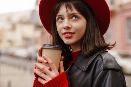 Close up of a smiling young stylish brunette white woman wearing hat and leather jacket with short hair standing on a street holding takeaway cupの写真素材