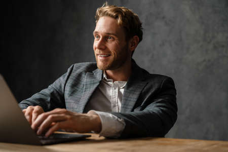 The portrait of smiling man typing on laptop while sitting at the table in the studio