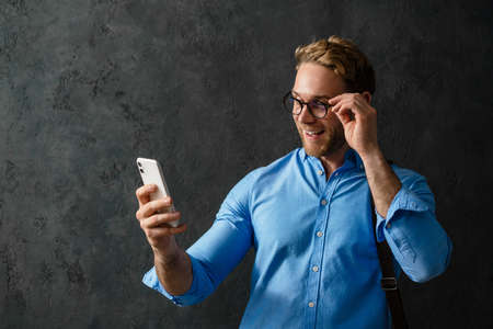 The portrait of smiling man in a blue shirt and glasses looking at the phone while standing in the...