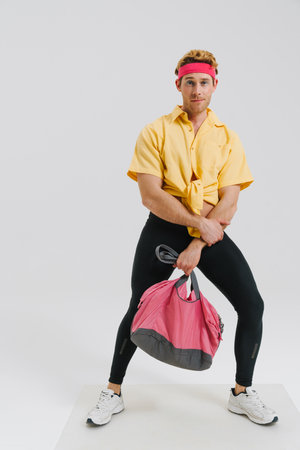 Young white gay man wearing headband standing over gray wall background carrying sportsbag
