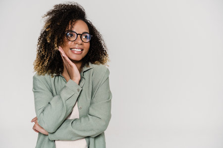 The smiling curly woman with glasses looking to the side and propping her face with her hand while standing in the studioの写真素材