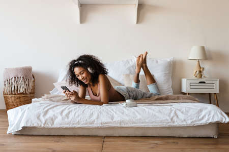 Smiling mid aged african woman listening to music with headphones while relaxing on bed at home,...