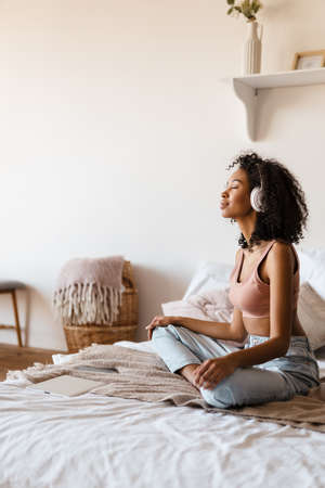 Smiling mid aged african woman listening to music with headphones while relaxing on bed at home, meditatingの写真素材