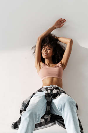 Young african woman wearing jeans smiling while lying on floor isolated over white wall