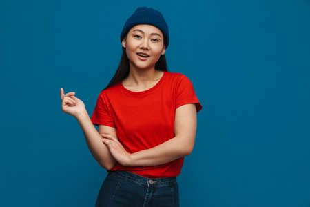 Asian teen girl in hat smiling and looking at camera isolated over blue backgroundの写真素材