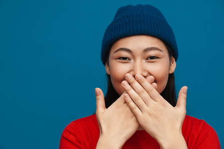 Happy young asian woman in casual clothes standing over blue wall background, covers mouth