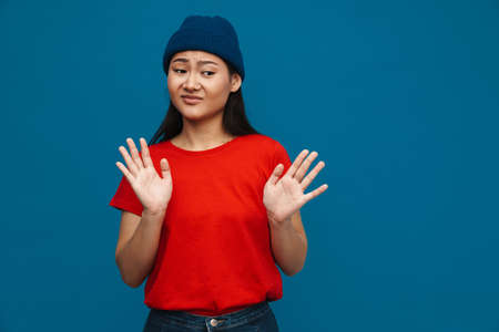 Displeased asian teen girl in hat showing stop gesture isolated over blue backgroundの写真素材
