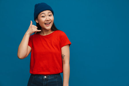 Asian teen girl in hat smiling and showing handset gesture isolated over blue backgroundの写真素材