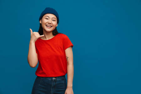 Asian teen girl in hat smiling and showing handset gesture isolated over blue backgroundの写真素材