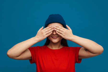 Happy young asian woman in casual clothes standing over blue wall background, covers face with handsの写真素材