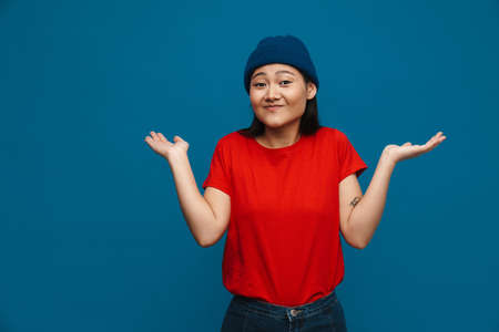 Smiling confused young asian woman in casual clothes standing over blue wall background, shrugging shouldersの写真素材