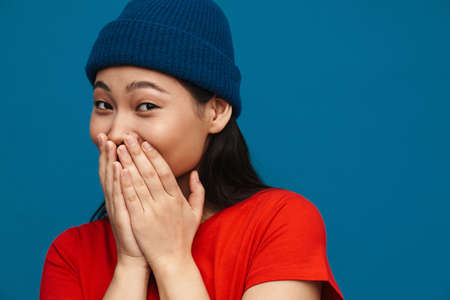 Asian teen girl in hat looking at camera and covering her mouth isolated over blue background