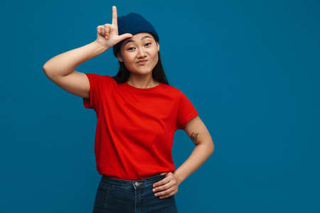 Asian teen girl in hat grimacing and pointing finger upward isolated over blue background