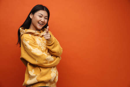 Asian teen girl wearing hoodie smiling and pointing finger at camera isolated over orange backgroundの写真素材