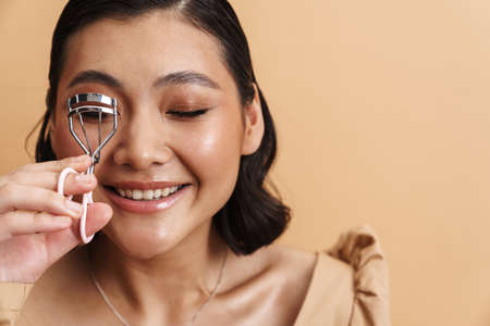 Young asian brunette woman smiling while using eyelash curler isolated over beige backgroundの写真素材