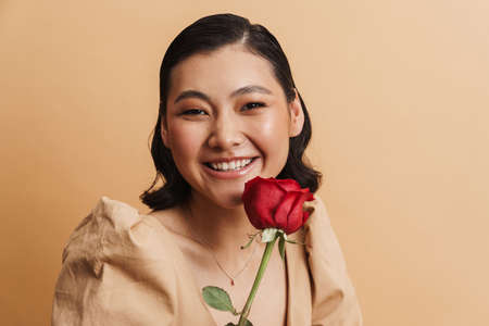 Young asian brunette woman smiling while posing with red rose isolated over beige backgroundの写真素材