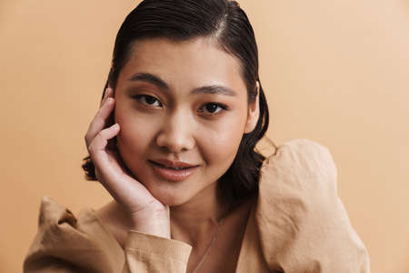 Young asian brunette woman posing and looking at camera isolated over beige background