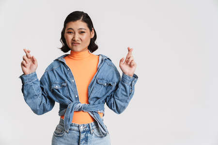 Young asian woman grimacing while holding fingers crossed for good luck isolated over white...