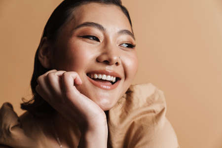 Young asian brunette woman laughing and looking aside isolated over beige background