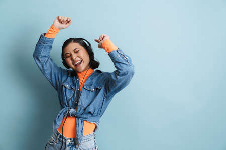 Young asian woman in headphones dancing and singing at camera isolated over blue backgroundの写真素材