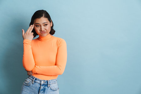 Portrait of a confused frowning casual brunette young asian woman standing over blue background showing gun gestureの写真素材