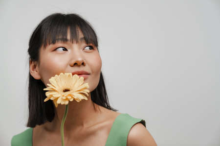 Young asian woman looking aside while posing with gerbera flower isolated over white backgroundの写真素材