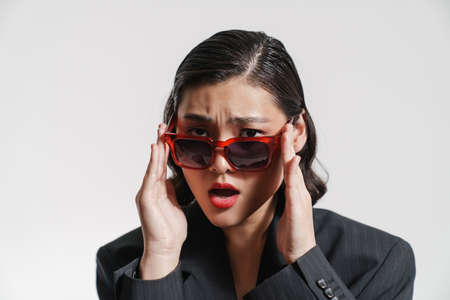 Brunette asian woman wearing jacket and sunglasses frowning at camera isolated over white backgroundの写真素材