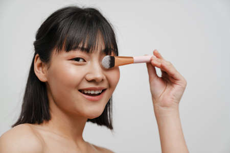 Asian shirtless woman using powder brush while doing makeup isolated over white backgroundの写真素材